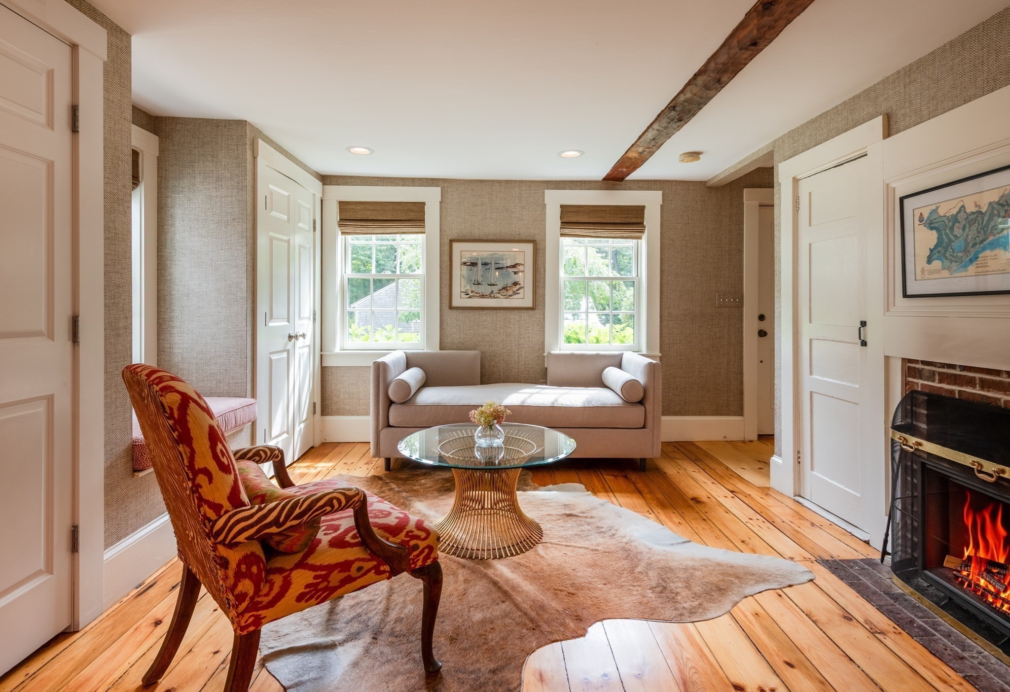 Living room with a patterned armchair, exposed ceiling beam, wood floors, and a lit brick fireplace.
