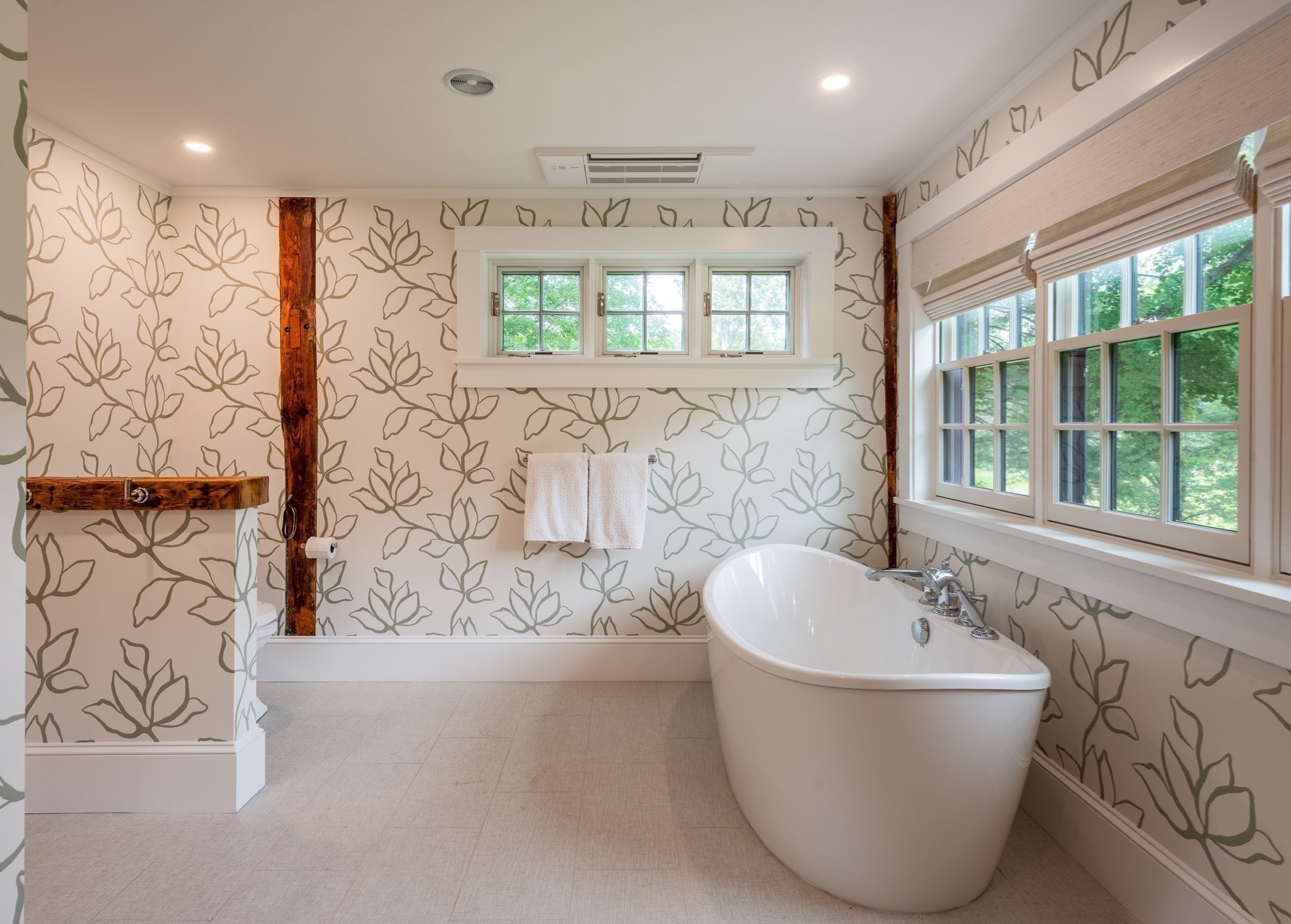 Bathroom with a freestanding soaking tub, botanical wallpaper, exposed wood beams, and large windows.