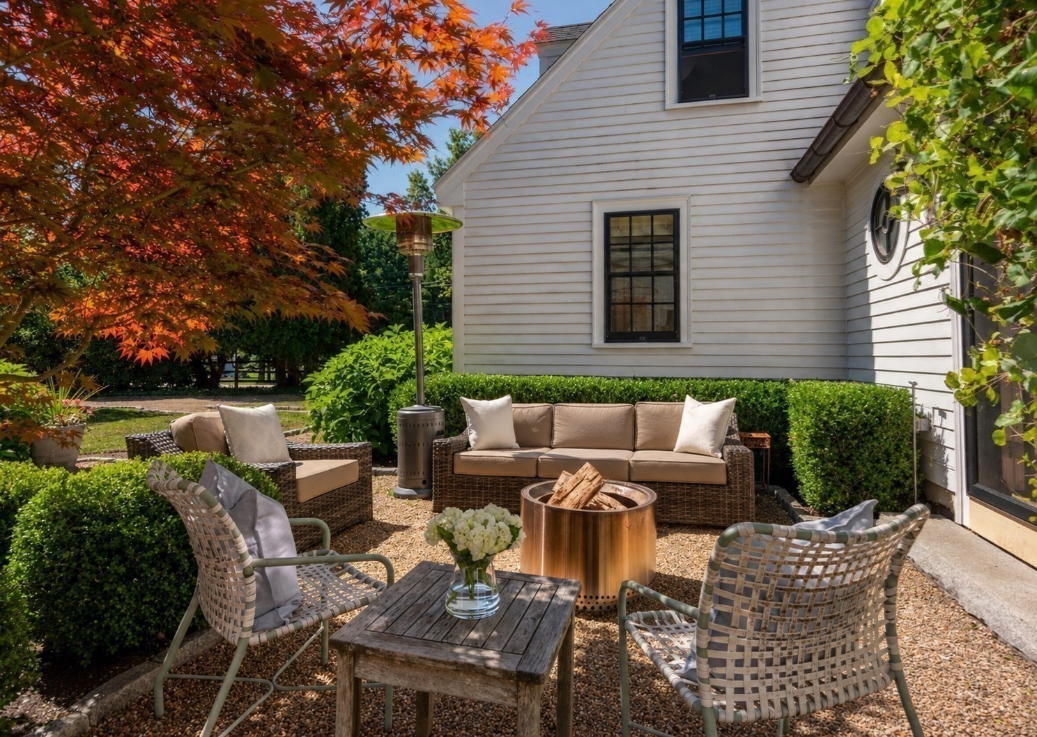 Furnished patio with wicker seating, a round fire pit, and red autumn foliage beside a white house.