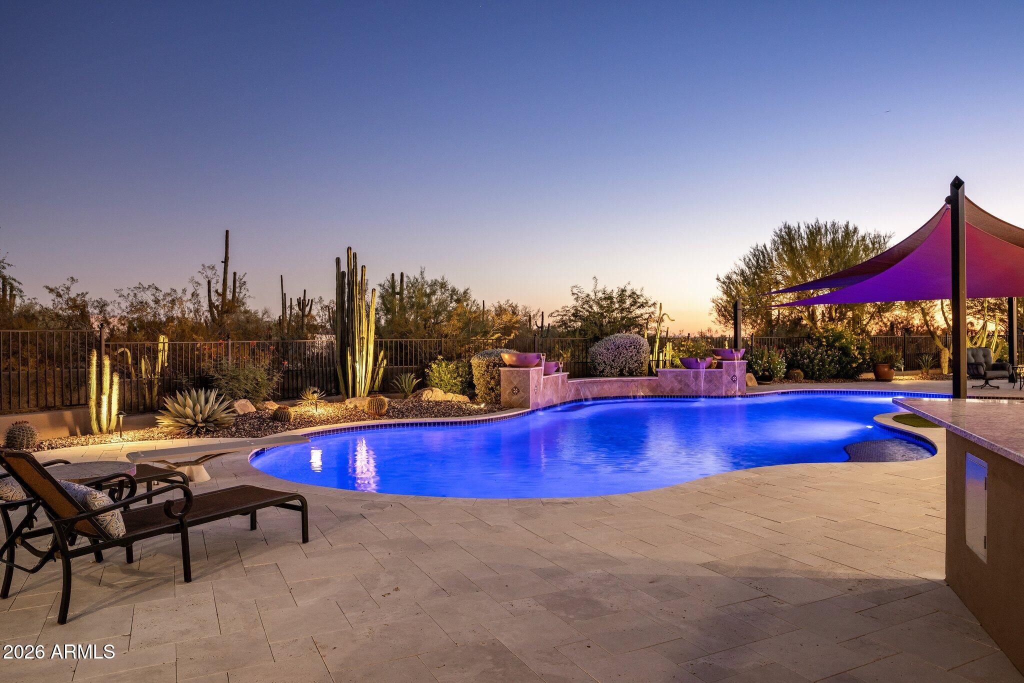 Curved backyard pool glows at dusk beside desert landscaping and a purple shade sail.