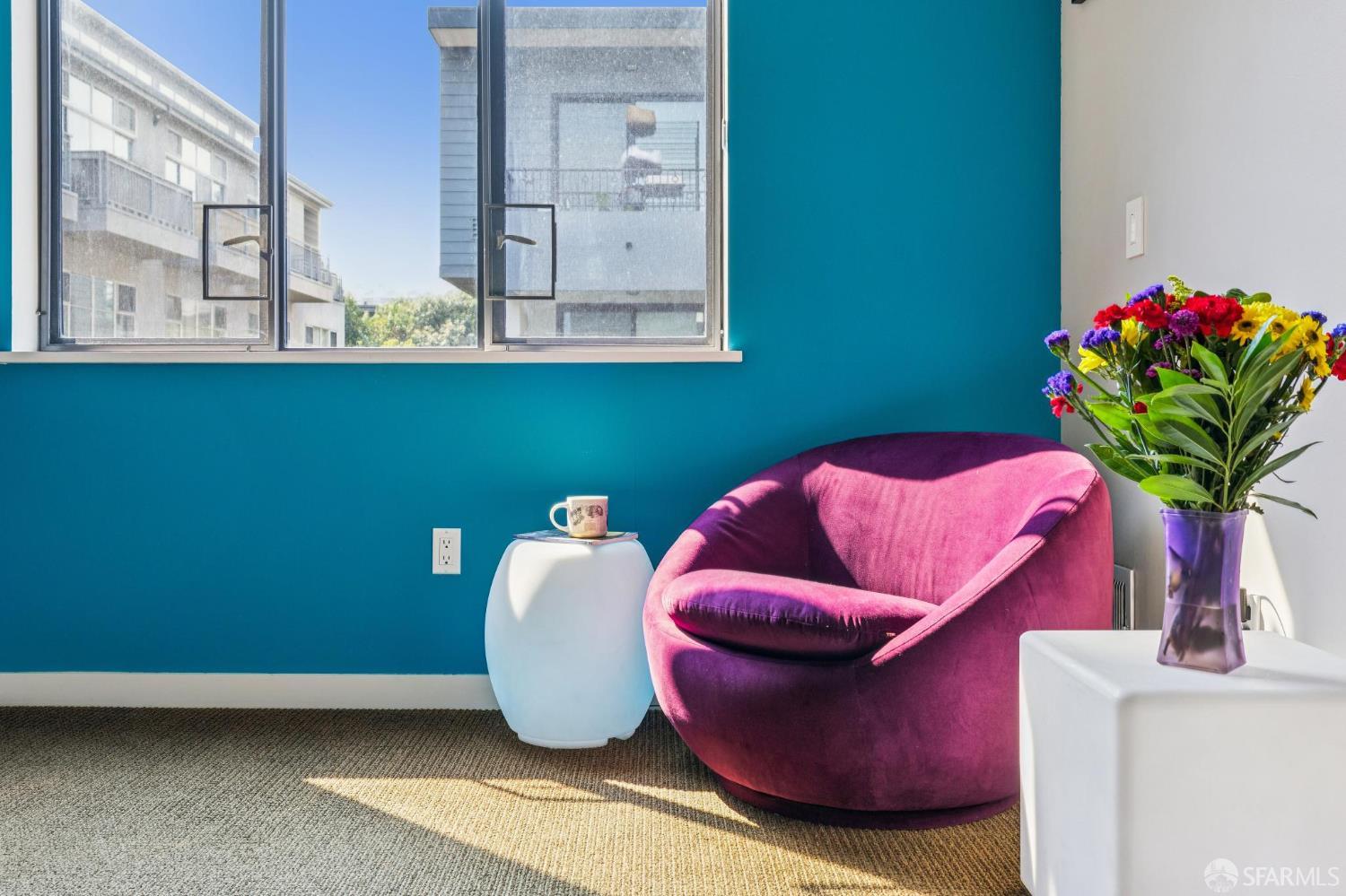 Furnished interior nook with a magenta chair, teal wall, sunlit window, and colorful flowers.