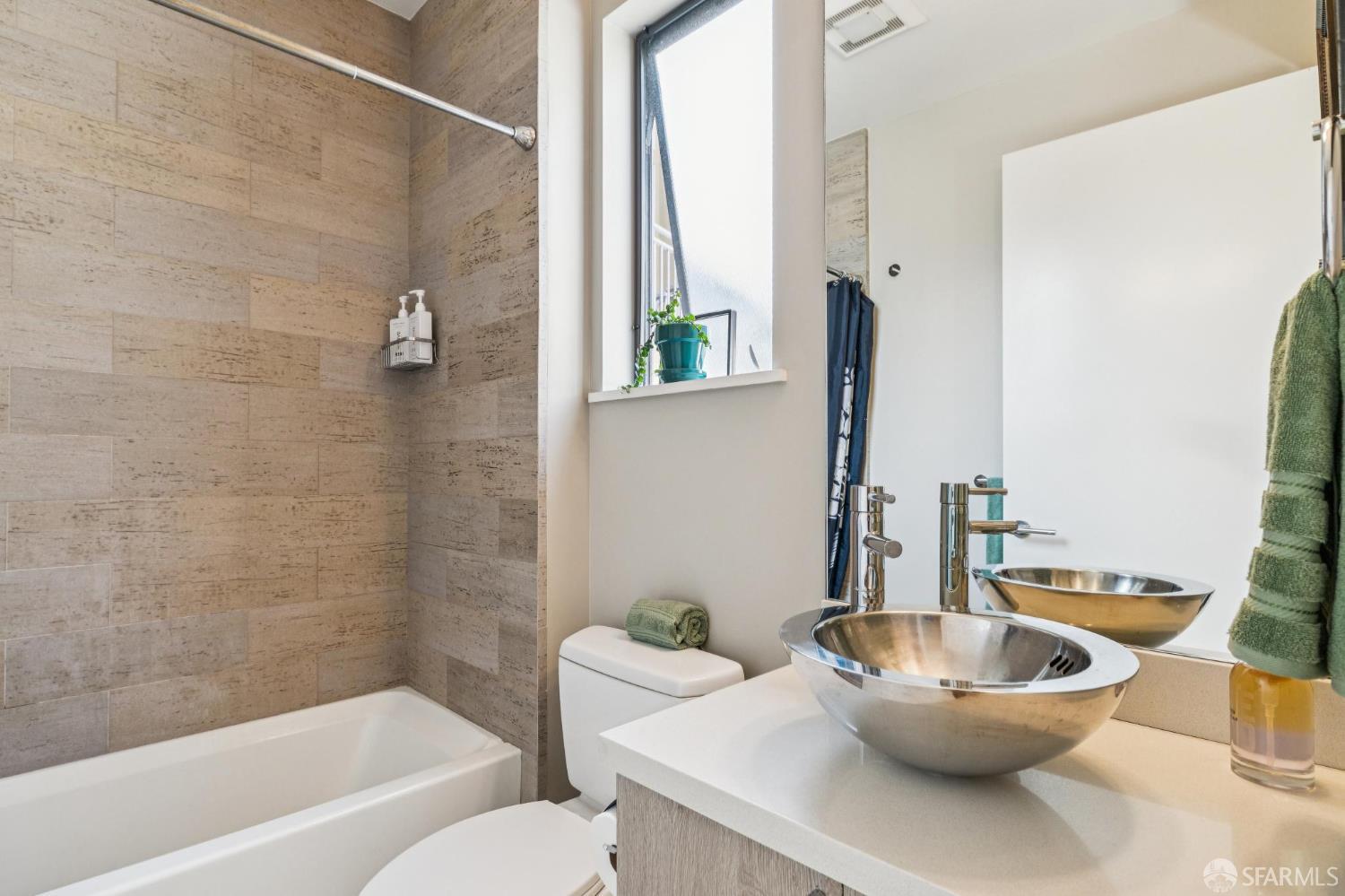 Bathroom with a beige stone-tile tub surround and polished metal vessel sink.