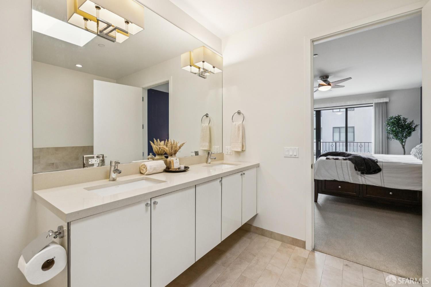 Bathroom with a long double-sink vanity and mirror, opening onto a bedroom with a bed and balcony doors.