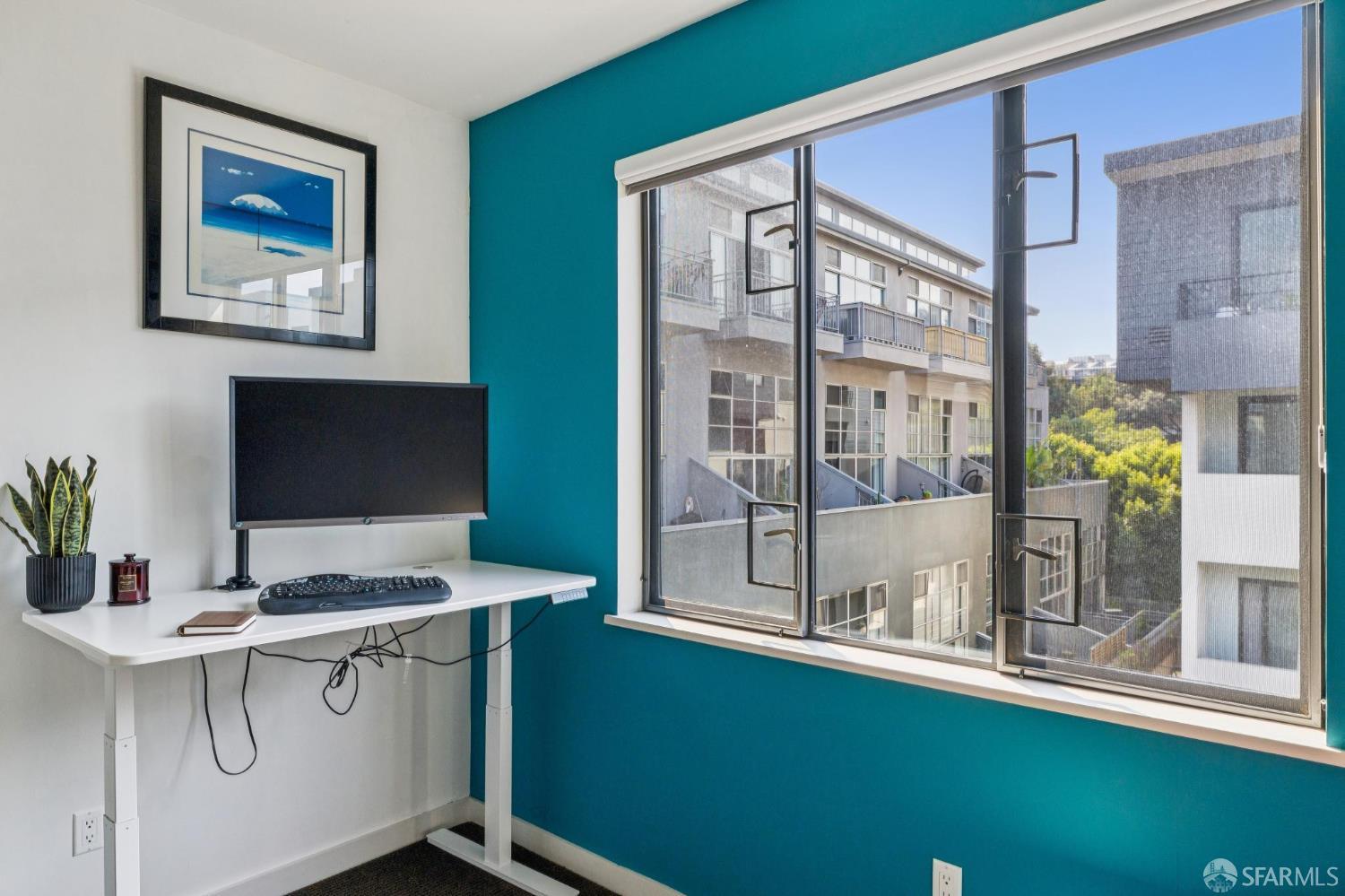 Home office with a standing desk beside a large window overlooking nearby buildings and trees.