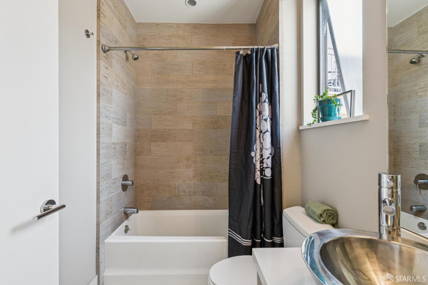 Bathroom with beige tiled tub surround, dark shower curtain, and a vessel sink beside a window.