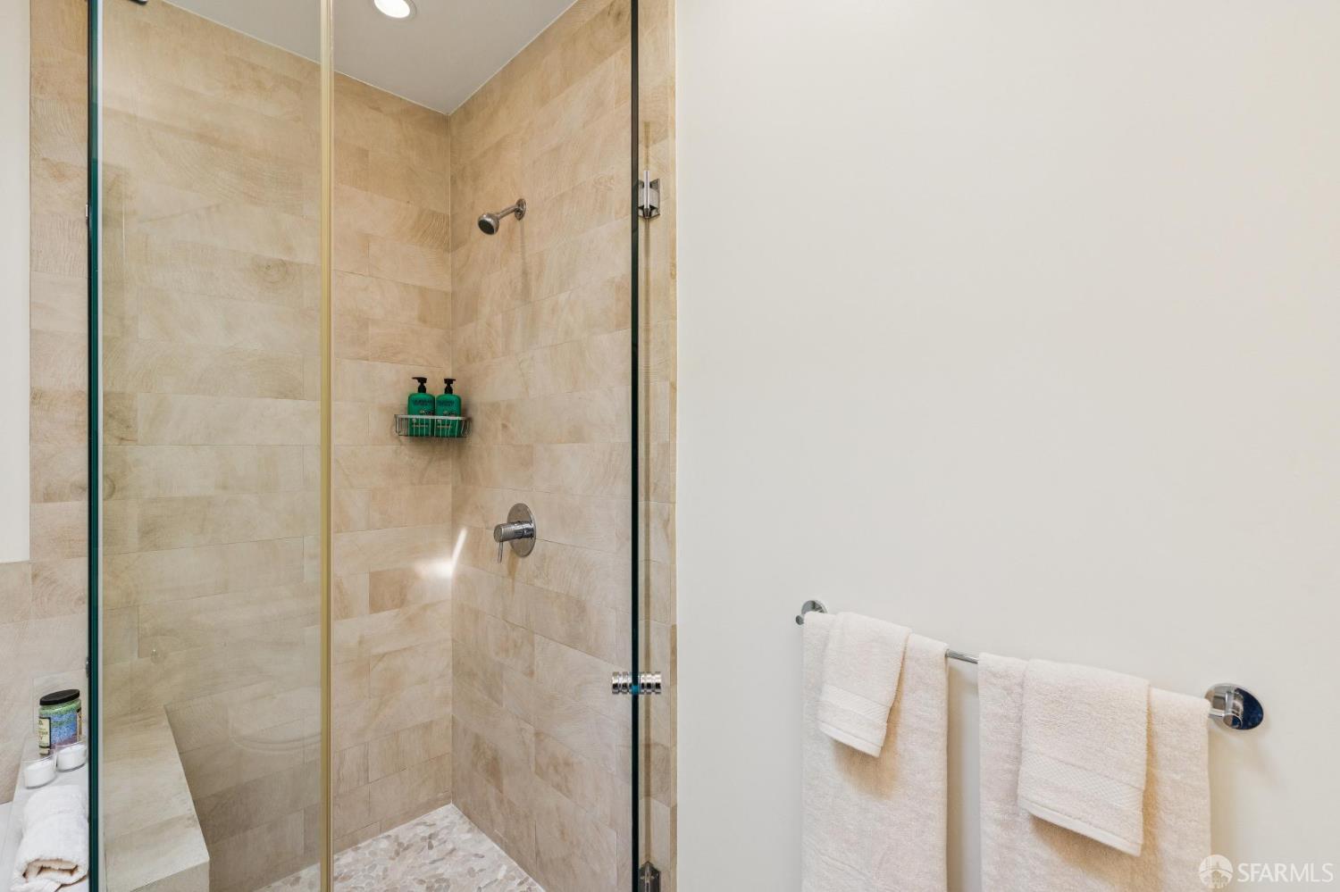 Bathroom shower with beige stone tile, a pebble floor, and white towels on a wall-mounted bar.