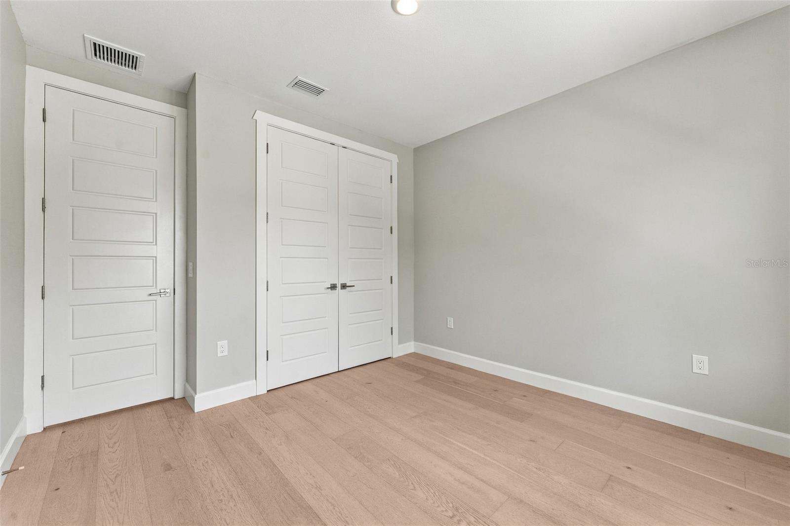 Empty bedroom with light wood flooring, pale gray walls, and white double closet doors.