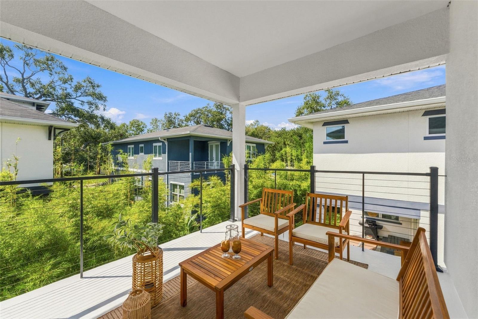 Covered deck furnished with wood lounge chairs and a coffee table, overlooking greenery and neighboring homes.