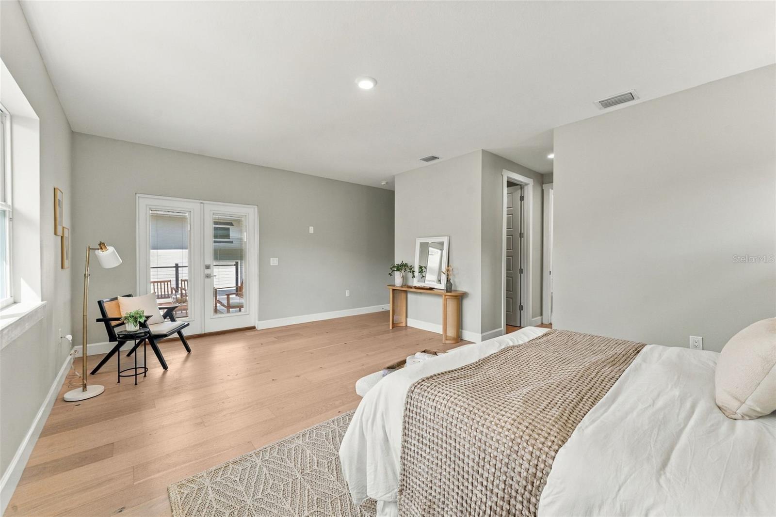 Staged bedroom with light wood floors, balcony doors, and a reading chair beside the window.