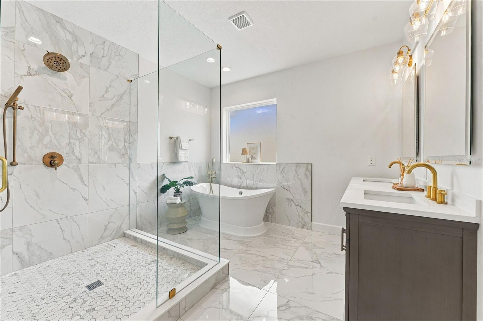 Bathroom with marble-look tile, a glass shower, freestanding tub, and dark vanity with gold fixtures.