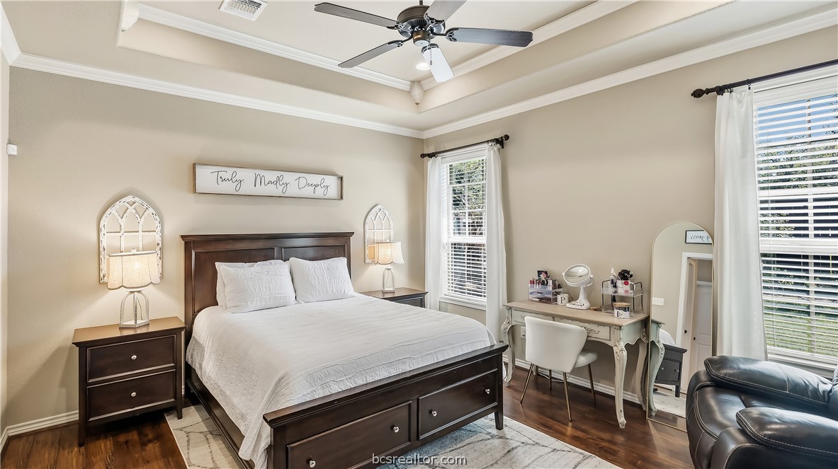 Bedroom with a dark wood bed, tray ceiling, ceiling fan, and dressing table beside tall windows.