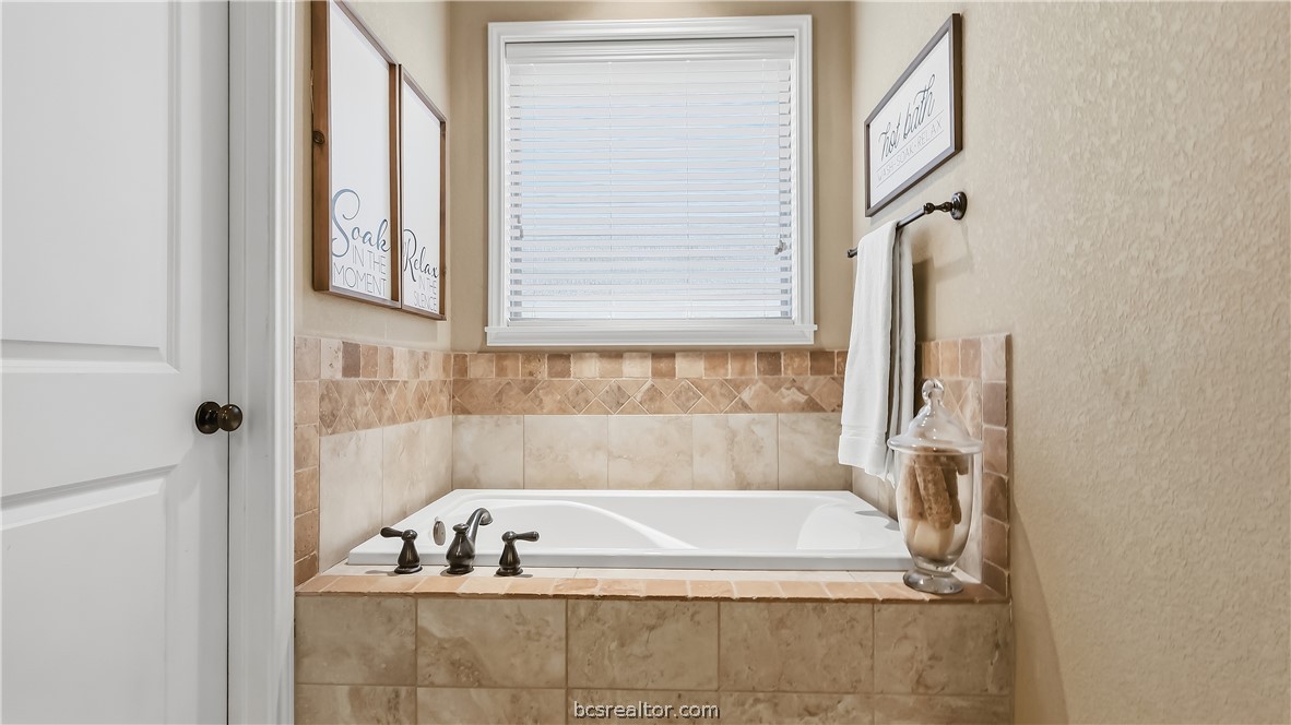 Bathroom soaking tub with tan tile surround, white blinds, and decorative wall signs.