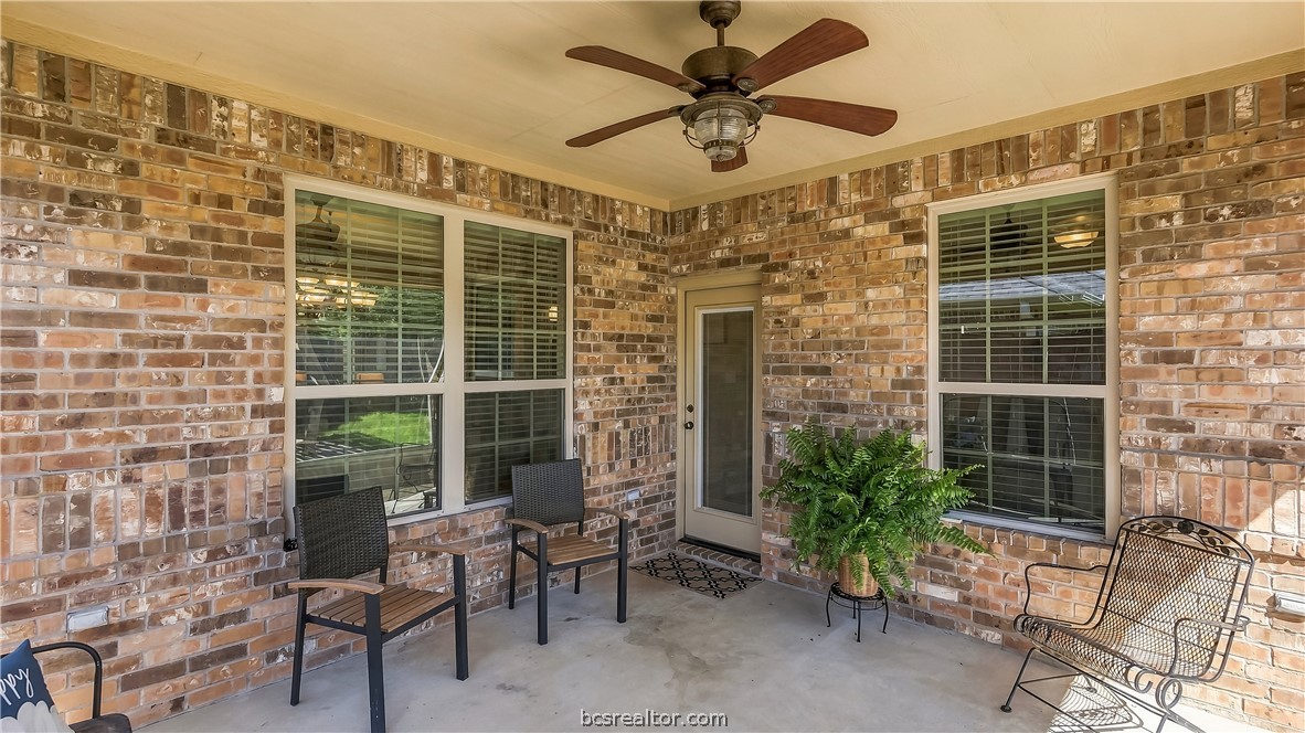 Covered brick patio with ceiling fan, two windows, glass door, chairs, and potted fern.
