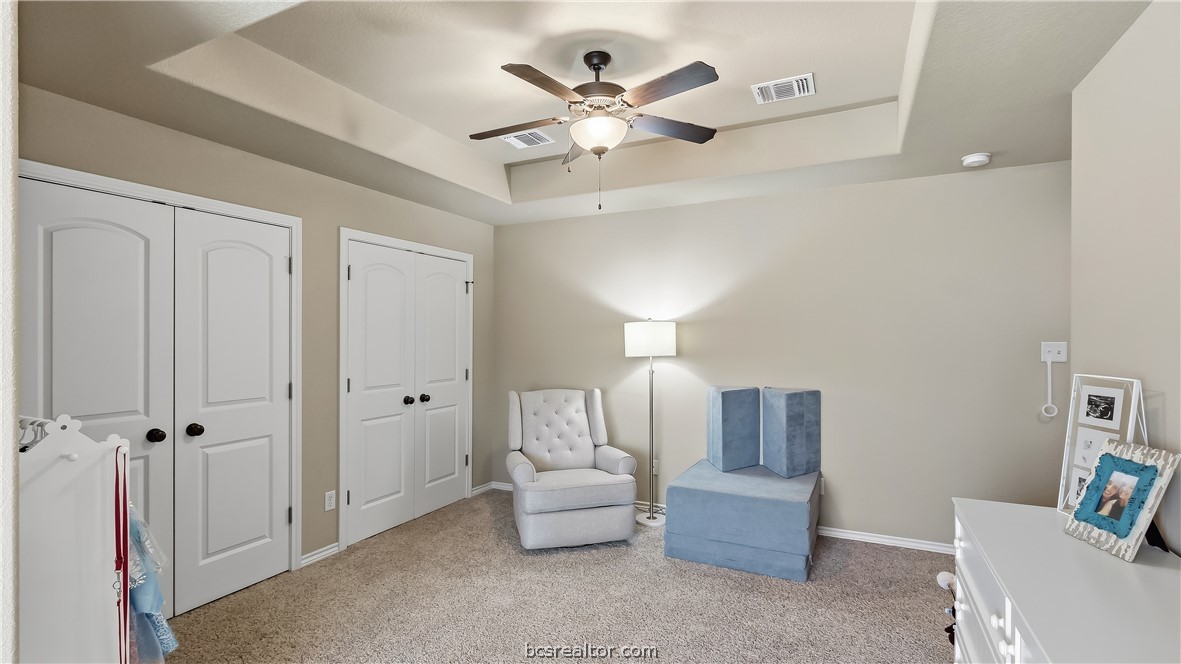 Furnished bedroom with a tray ceiling, ceiling fan, closet doors, and upholstered seating.