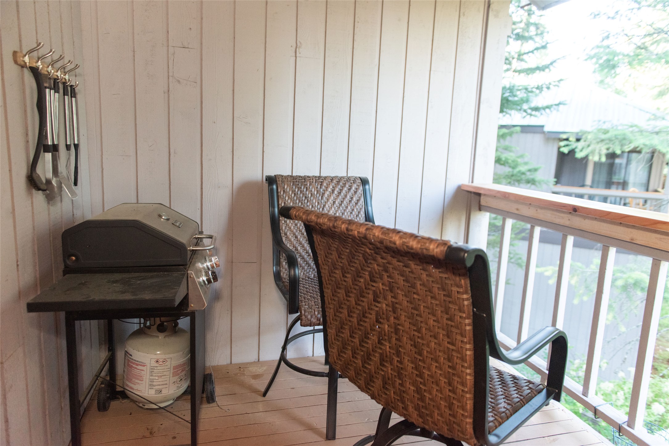 Condo balcony with woven chairs and a propane grill beside a leafy view.