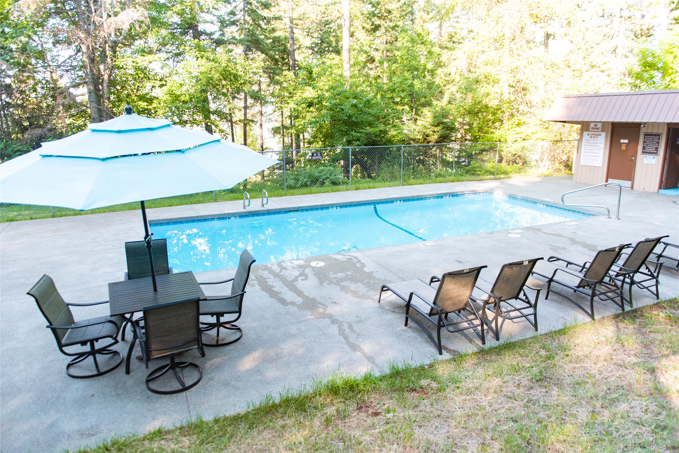 Outdoor pool with a large umbrella, patio table, and lounge chairs beside a wooded fence.