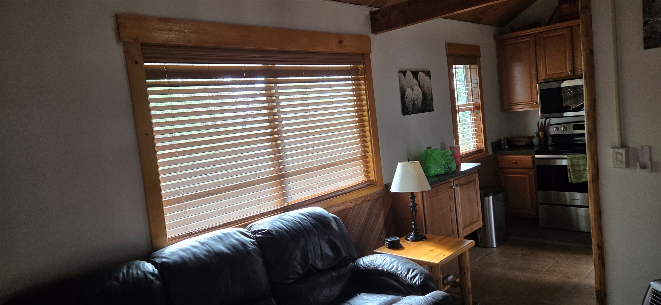 Condo living area with dark leather sofa beneath a wide blinded window and a compact wood-cabinet kitchen.