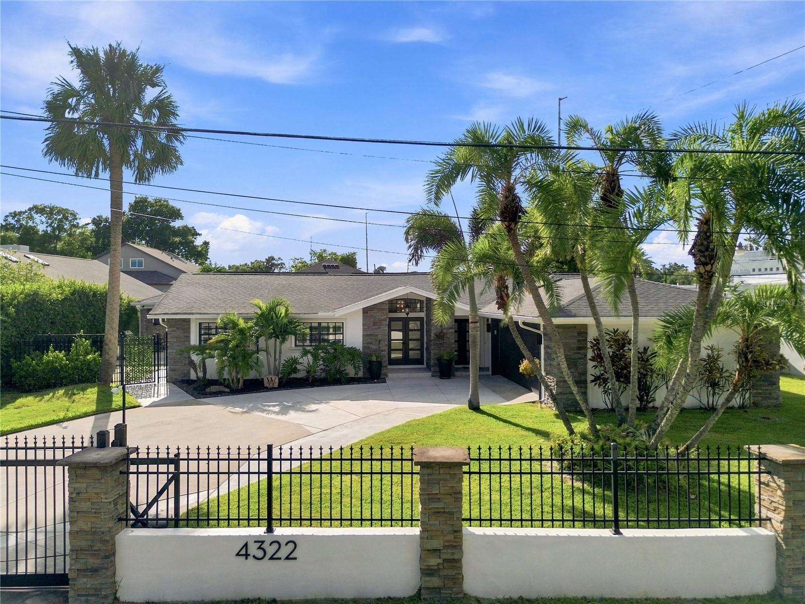 Single-story home behind a black metal gate, with stone accents, a broad driveway, and palm trees.