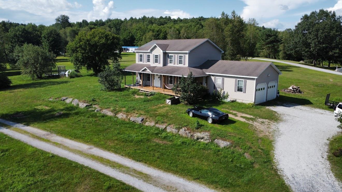Aerial view of a gray two-story home with an attached garage, covered porch, and wooded lawn.