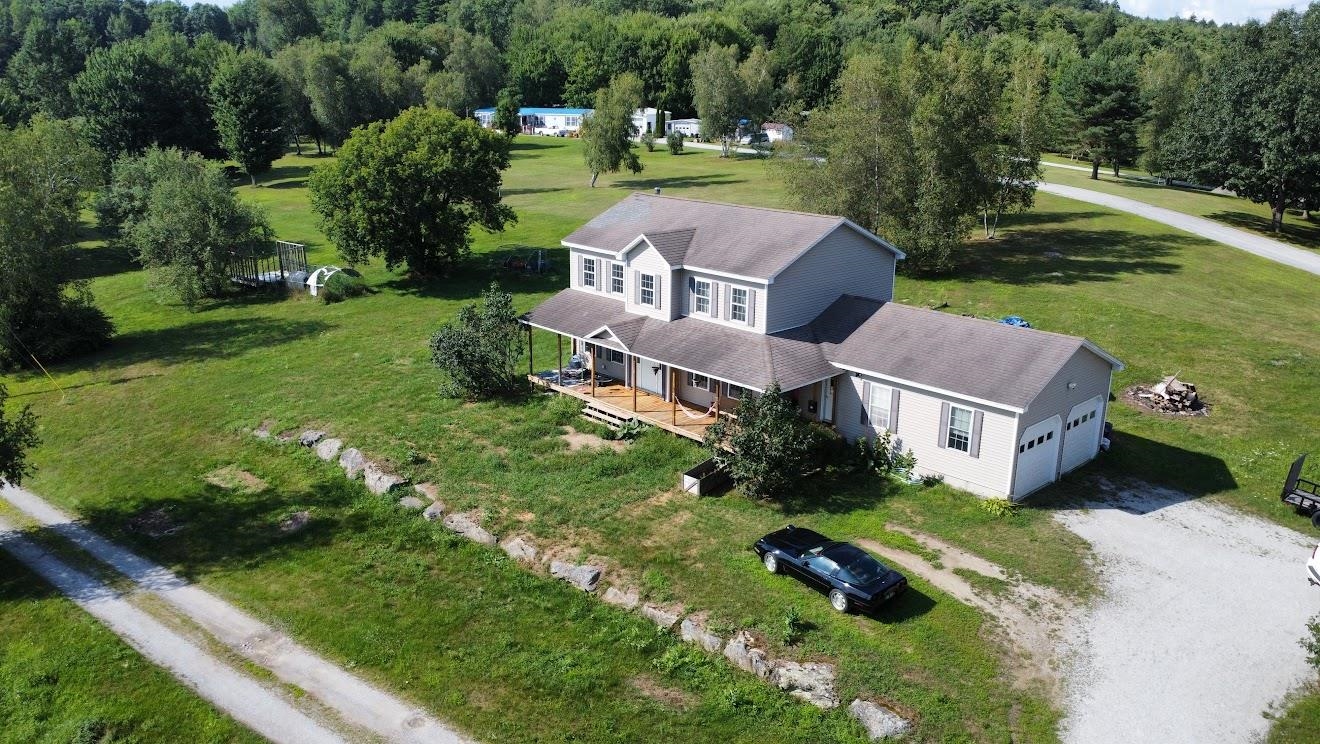 Aerial view of a gray two-story house with a covered porch, attached garage, and broad tree-lined lawn.