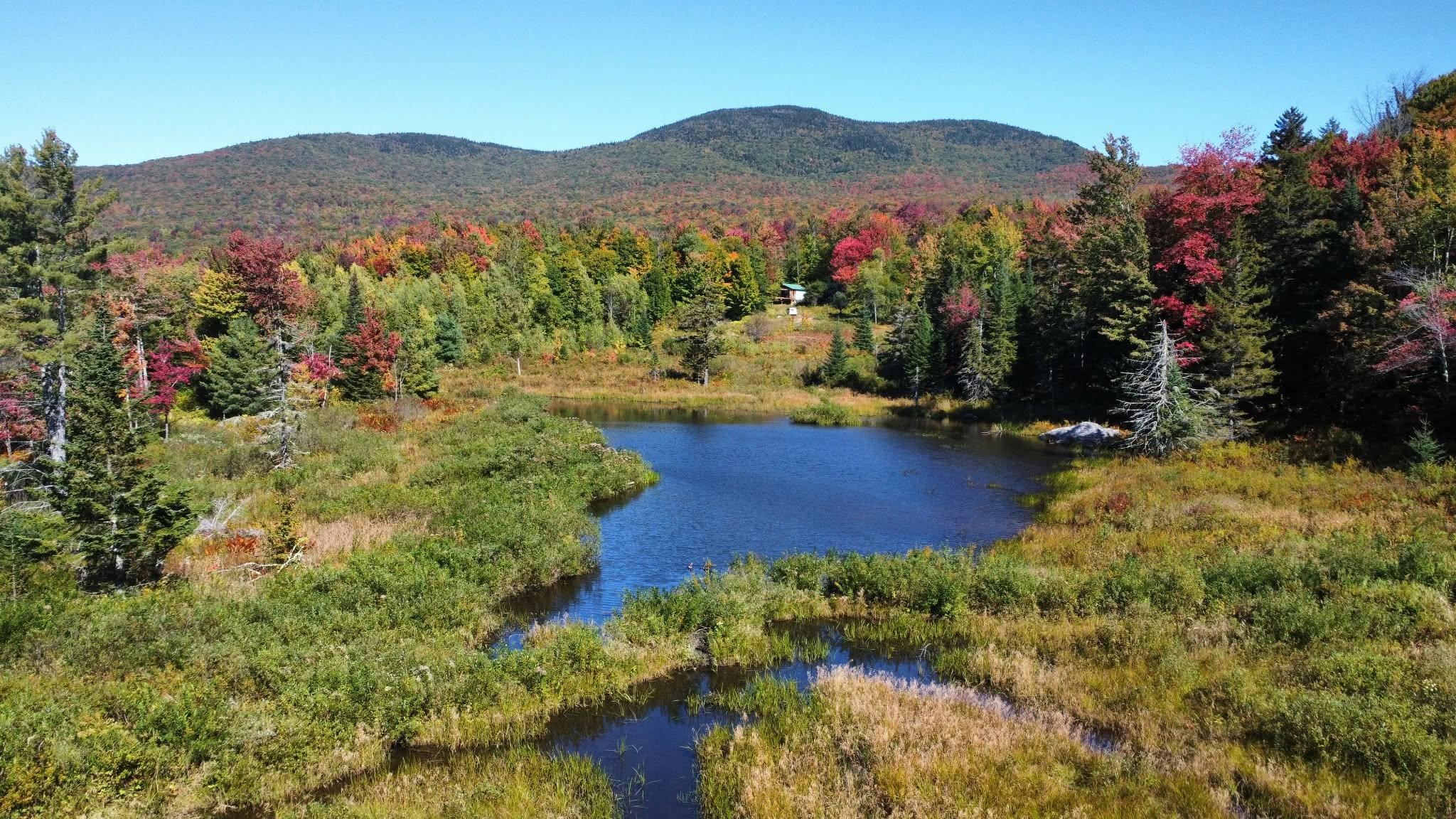 Aerial view of a pond amid autumn foliage, with forested mountains and a small structure beyond.