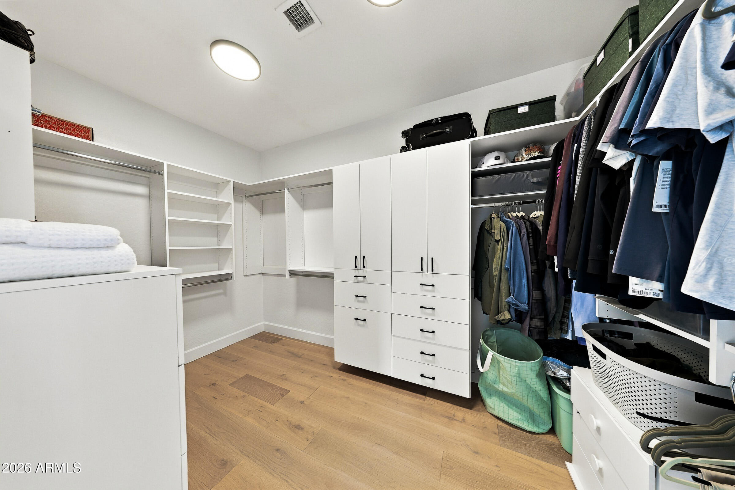 Walk-in closet with white built-in storage, hanging clothes, and warm wood flooring.