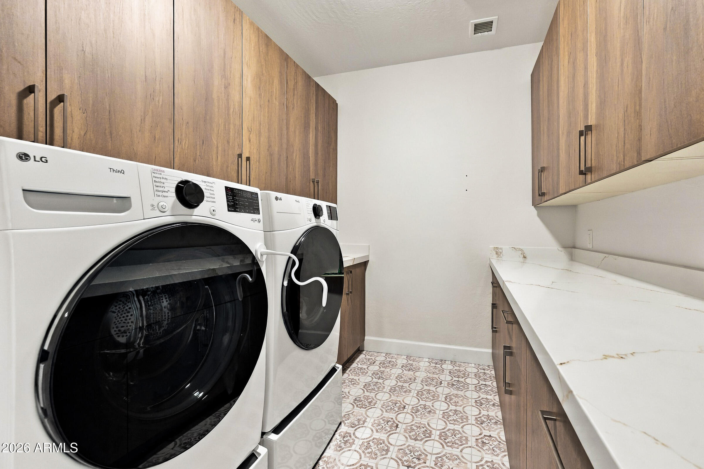 Laundry room with front-loading washer and dryer, wood cabinets, white countertop, and patterned tile floor.