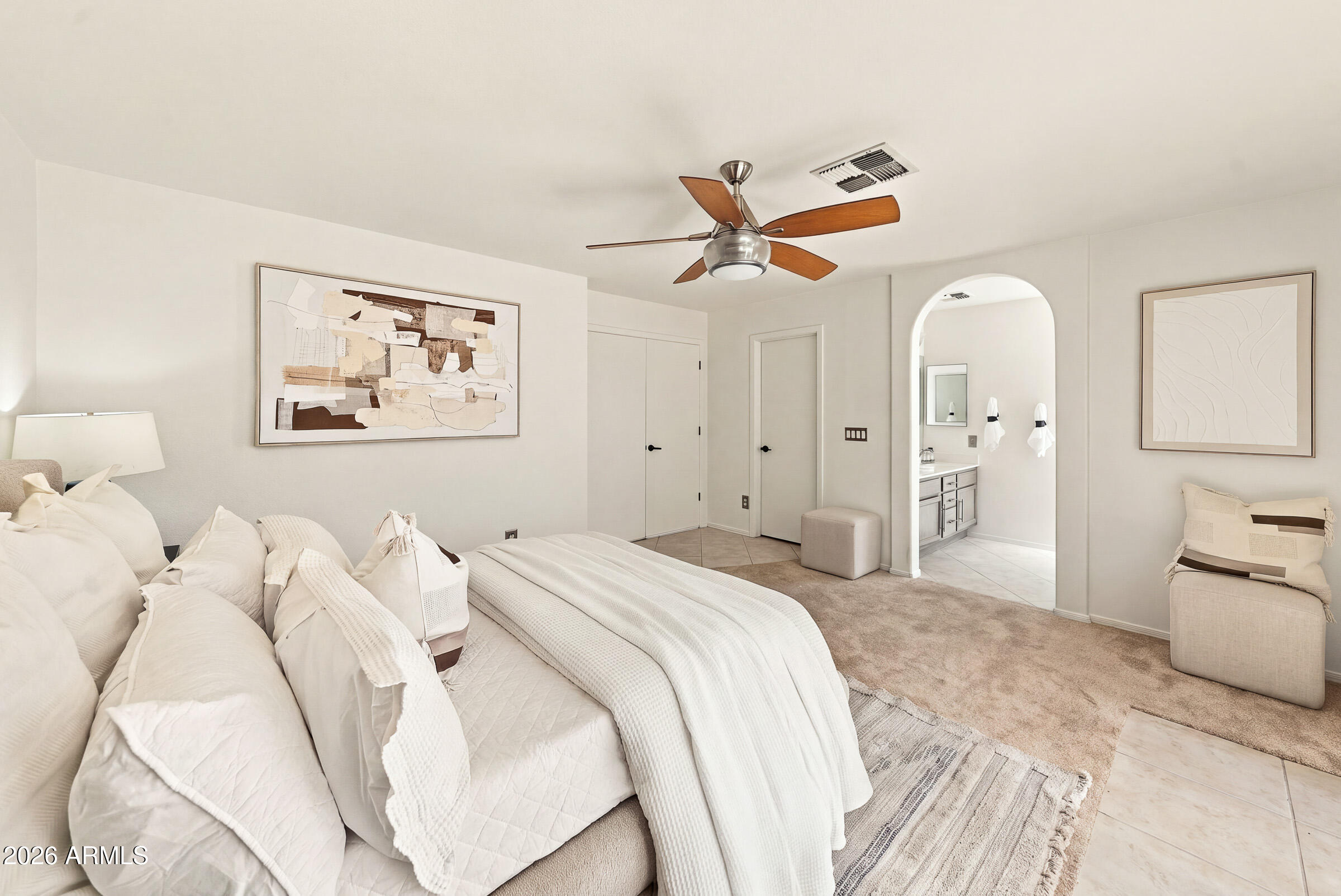 Staged bedroom with a large white bed, wood-blade ceiling fan, and arched opening to the bathroom.