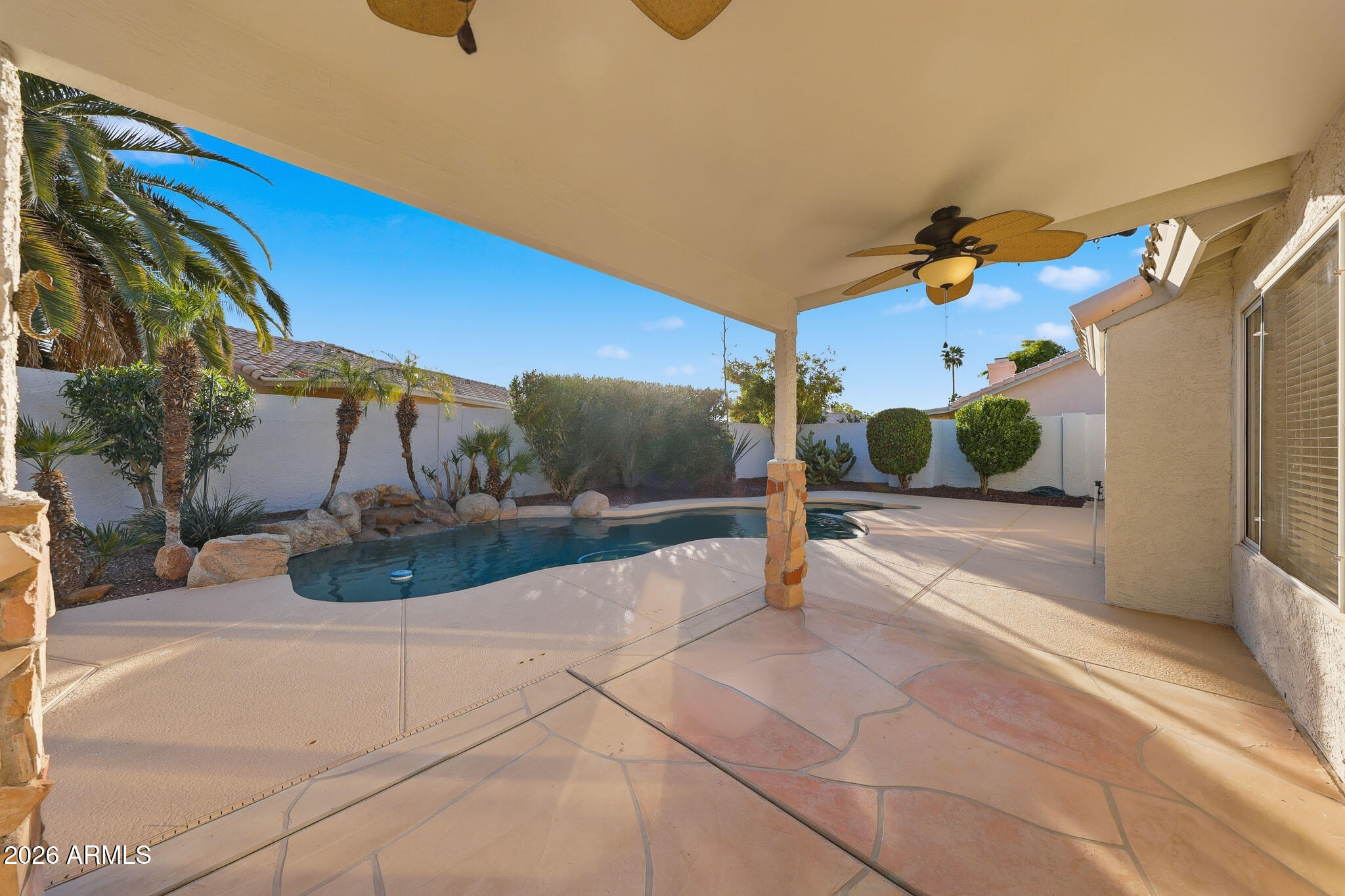 Backyard pool and paved patio beneath a covered area with a ceiling fan and desert landscaping.