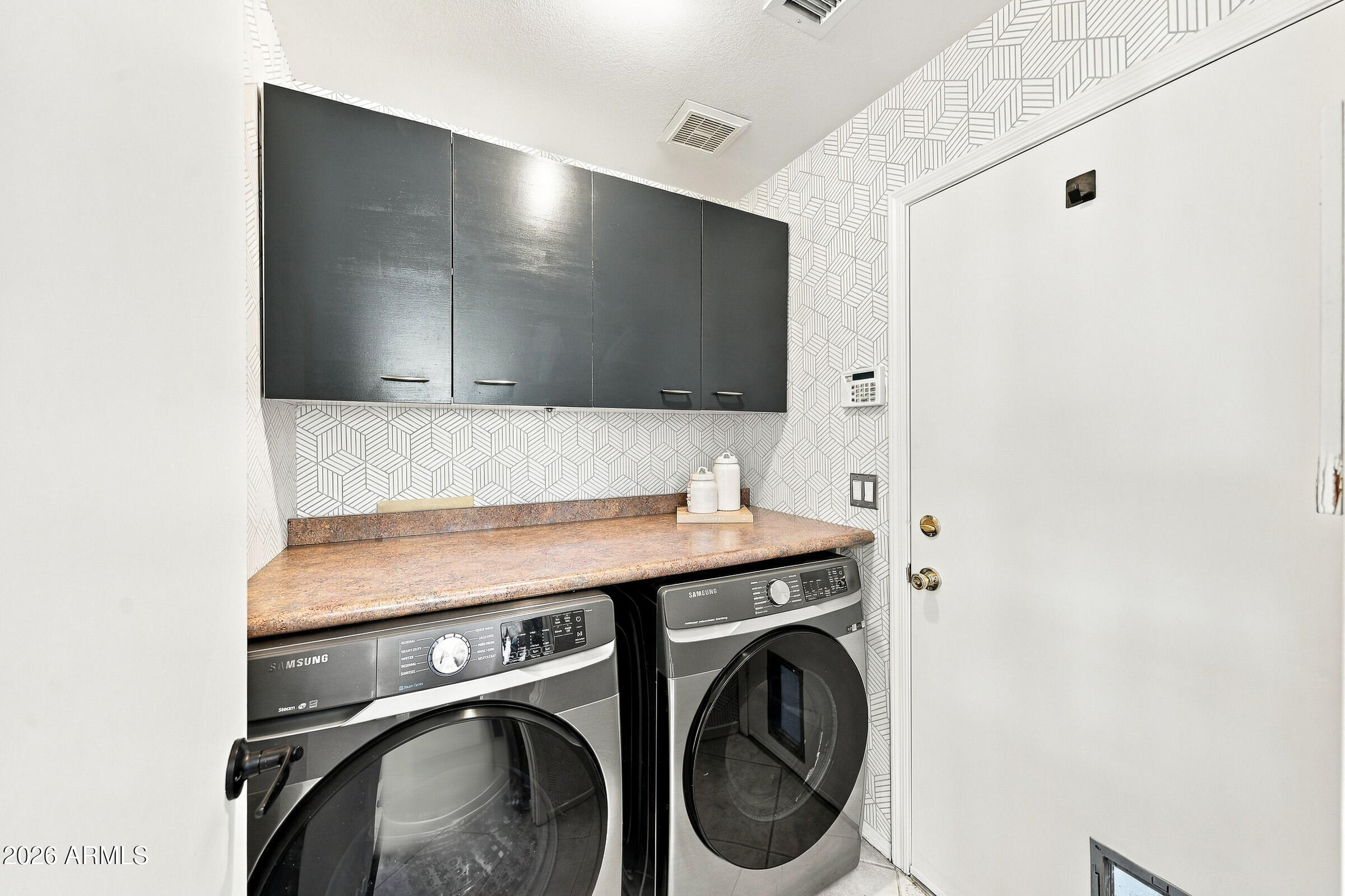 Laundry room with front-loading washer and dryer, dark cabinets, and geometric patterned wallpaper.