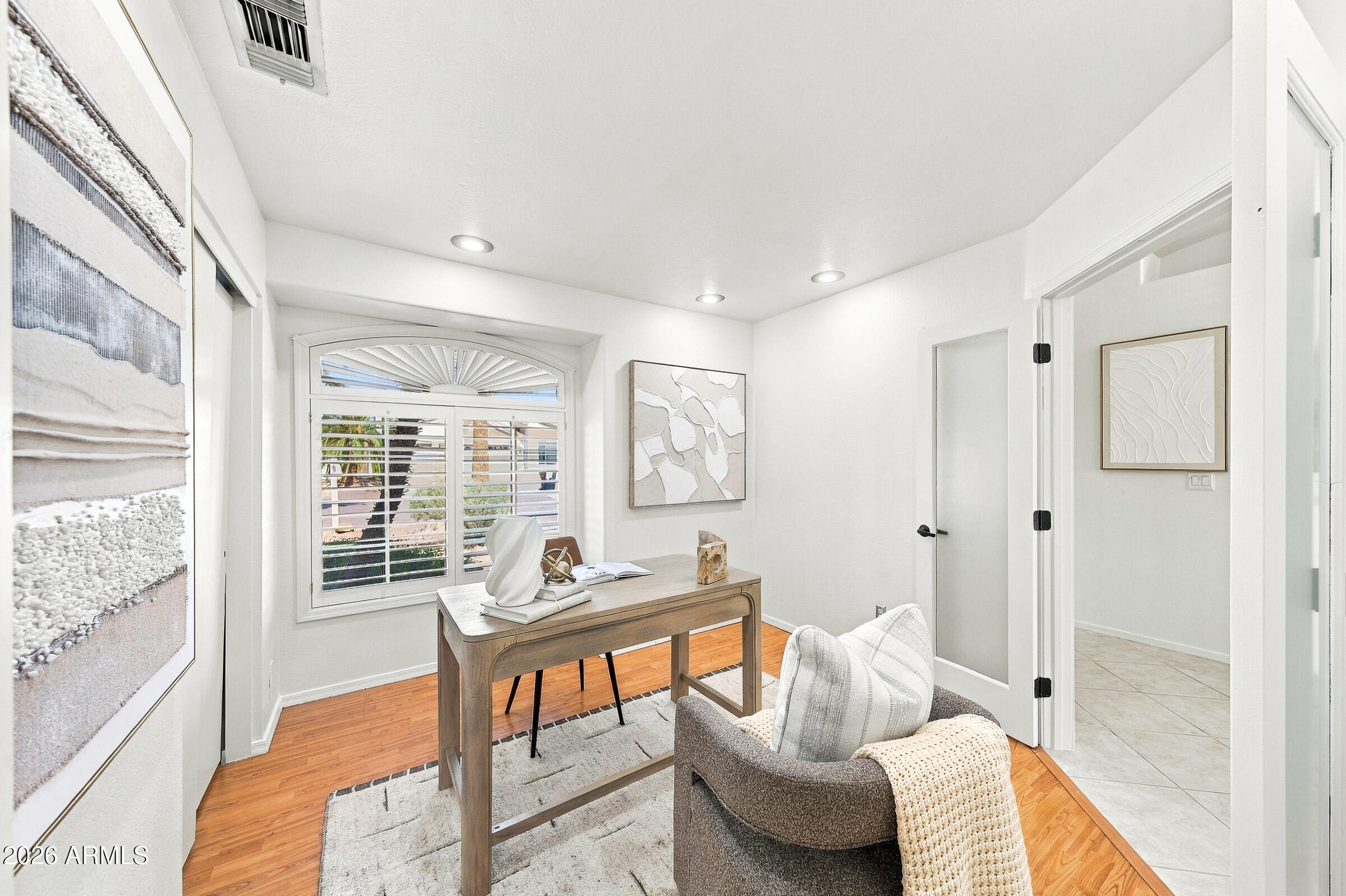 Furnished home office with a wooden desk, cushioned chair, and arched shuttered window.