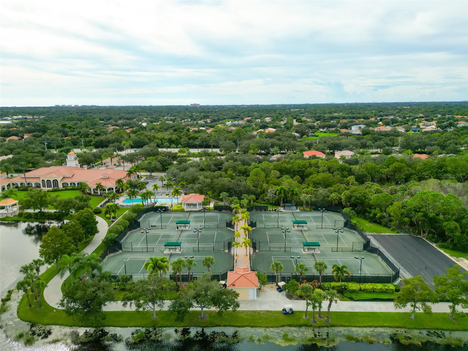 Aerial view of community tennis courts, pool, clubhouse, and pond surrounded by trees.