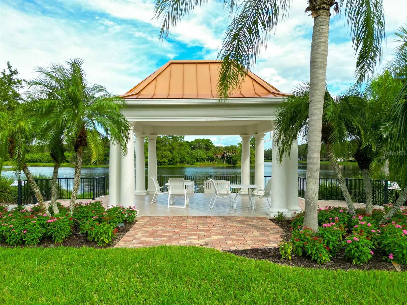 Waterfront pavilion with white columns, outdoor dining furniture, and palm trees beside a lake.