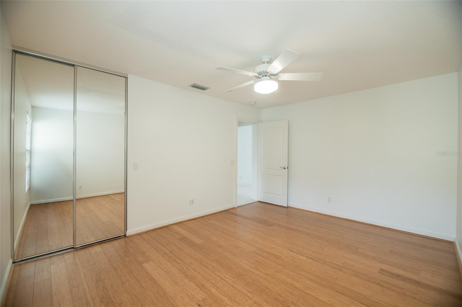 Empty bedroom with wood flooring, mirrored closet doors, and a ceiling fan with light.
