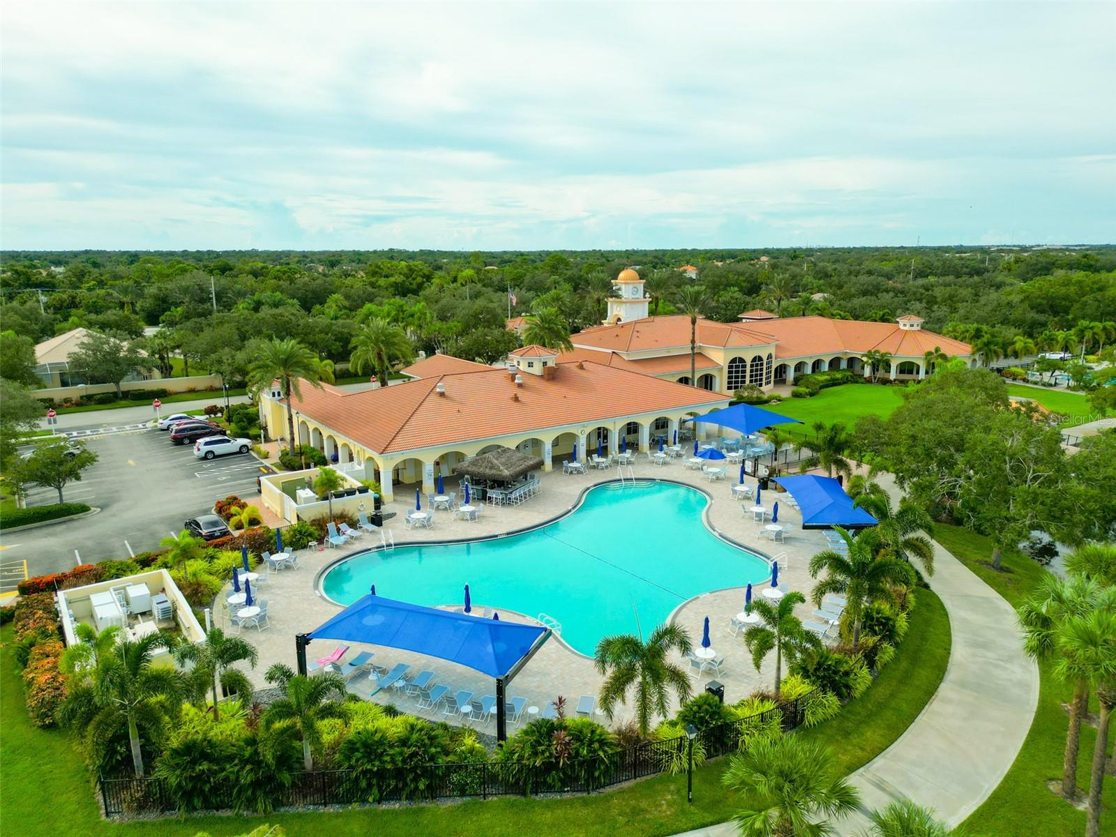 Aerial view of a community pool and clubhouse with orange tile roofs and blue shade canopies.