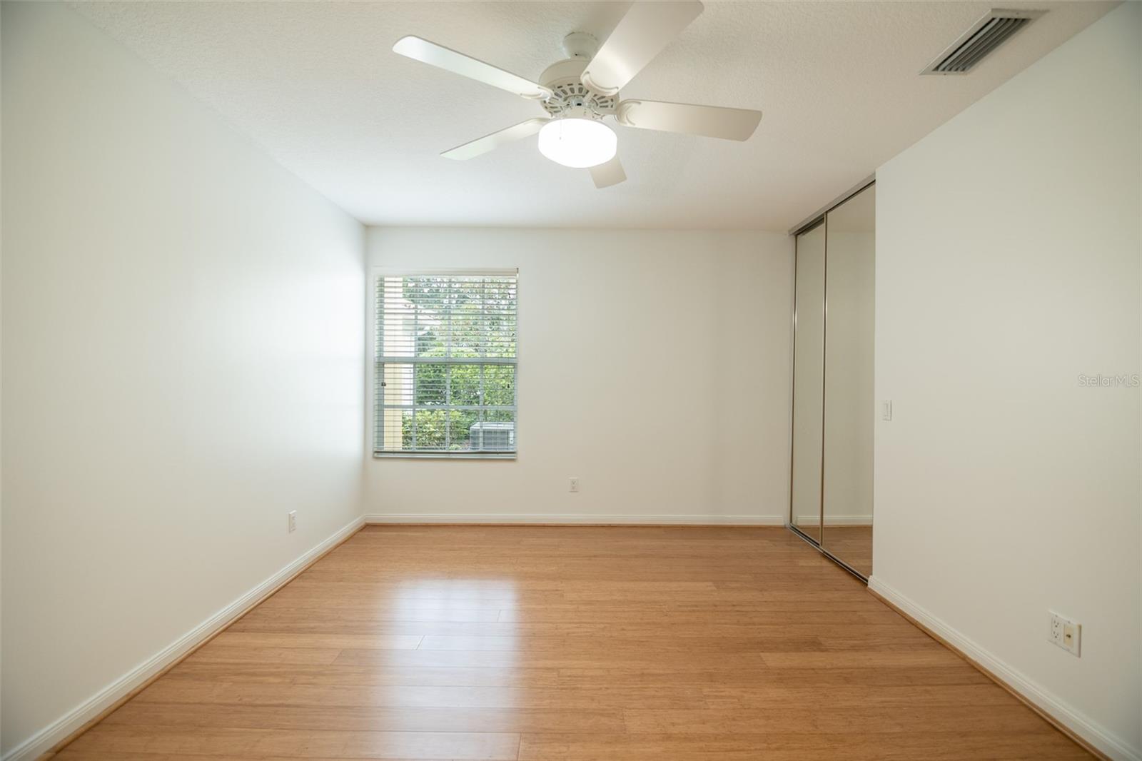 Empty bedroom with wood flooring, mirrored closet doors, and a ceiling fan.