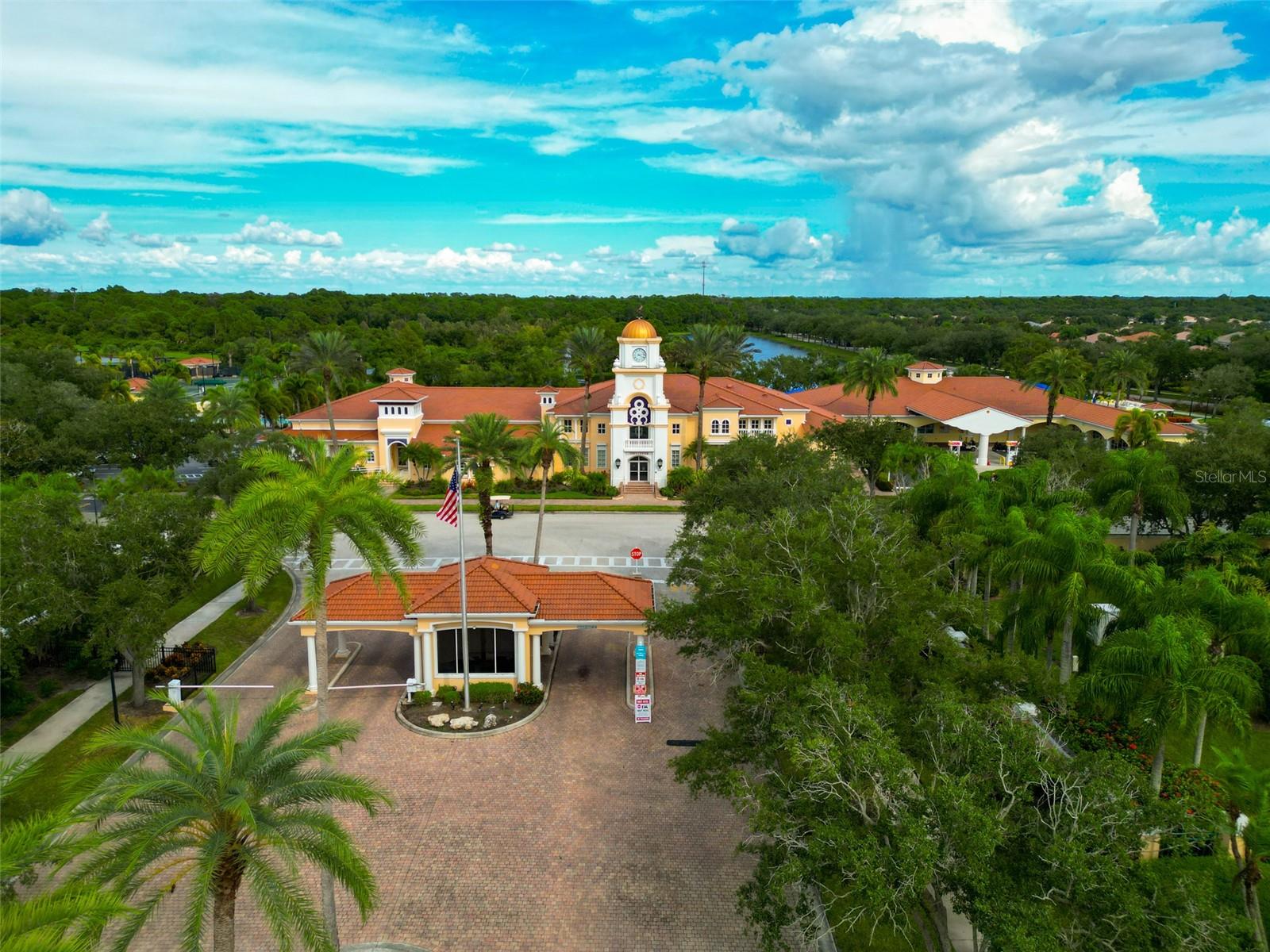 Community clubhouse with a central clock tower, terracotta roofs, and palm-lined entrance drive.