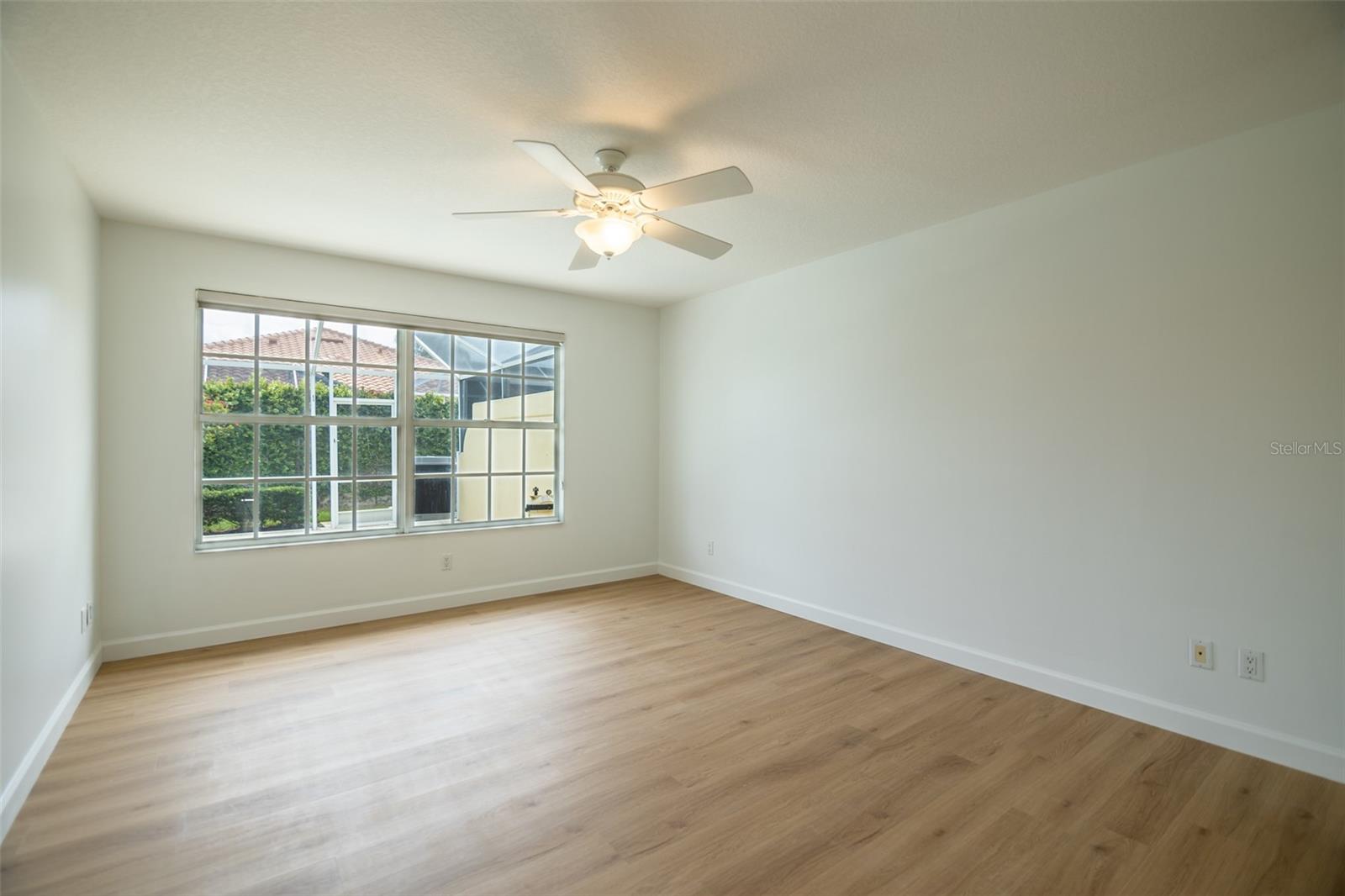 Empty room with light wood floors, a ceiling fan, and a large window facing greenery.