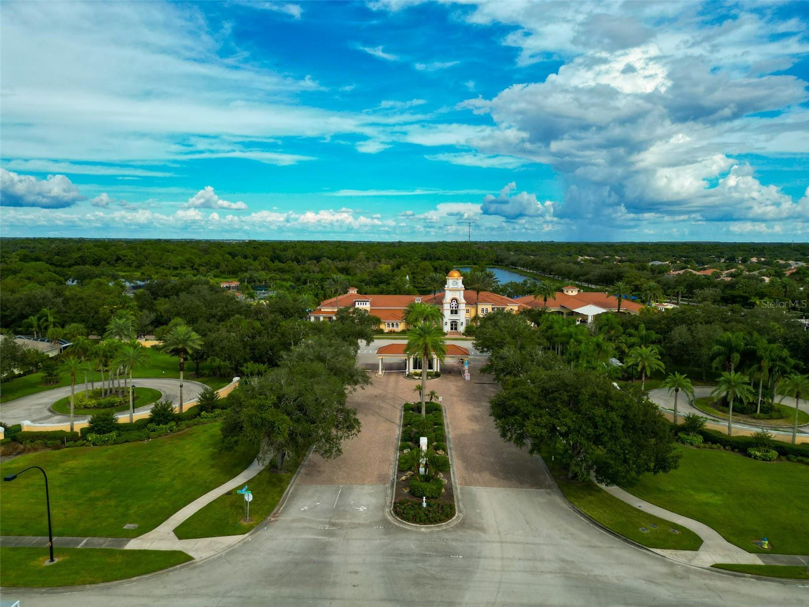 Aerial view of a Mediterranean-style clubhouse with a central tower, palm-lined circular drive, and wooded grounds.