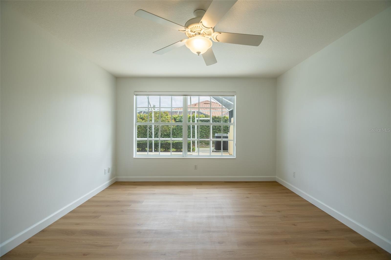 Empty room with wood-look flooring, a ceiling fan, and a wide window overlooking greenery.