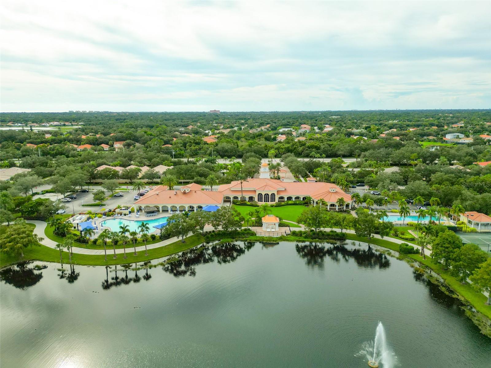 Aerial view of a community clubhouse, pools, tennis courts, and fountain pond.