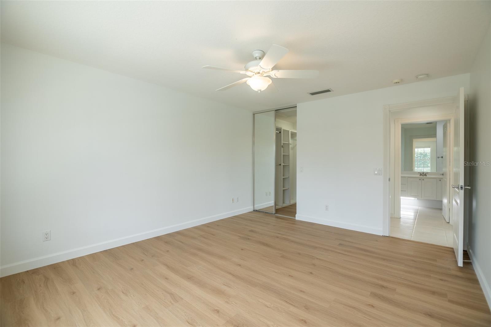 Vacant bedroom with light wood floors, mirrored closet doors, and an adjoining bathroom.