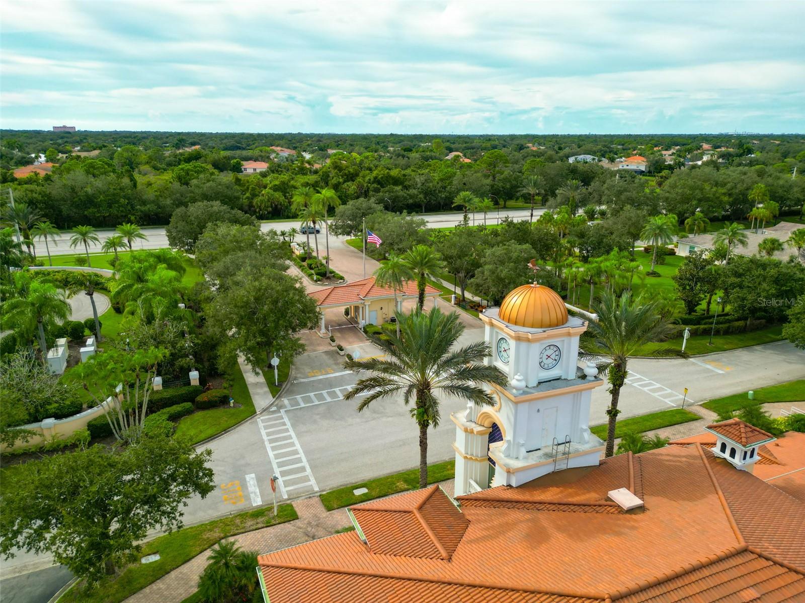 Aerial view of a residential community entrance with a gold-domed clock tower, terracotta roofs, and palm-lined grounds.