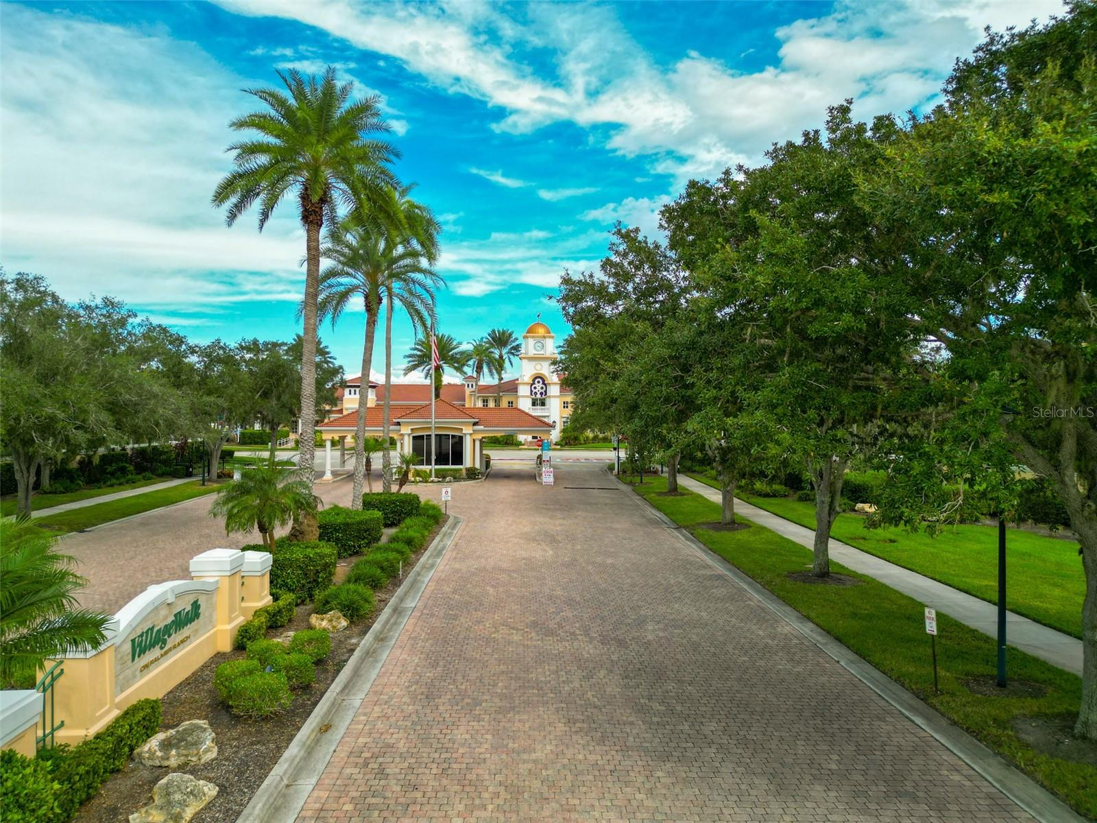 Landscaped residential community entrance with palm trees, a brick-paved drive, and a clock-tower building.