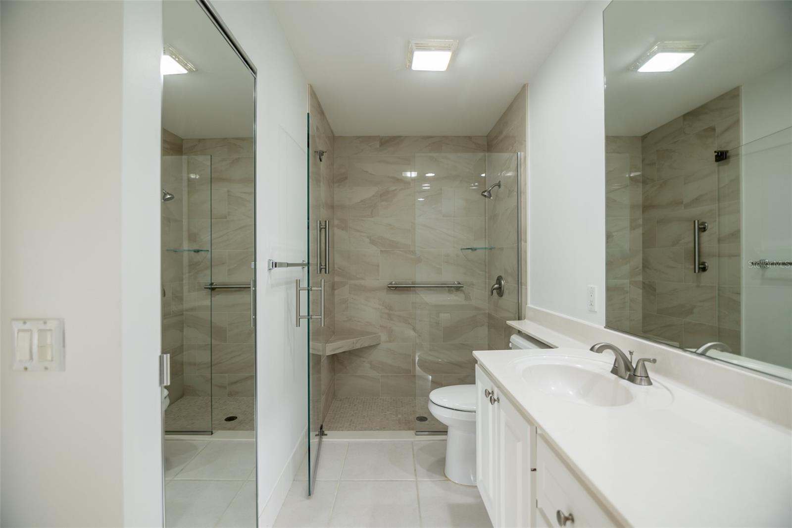 Bathroom with a glass walk-in shower, marble-look tile, built-in bench, grab bars, and white vanity.