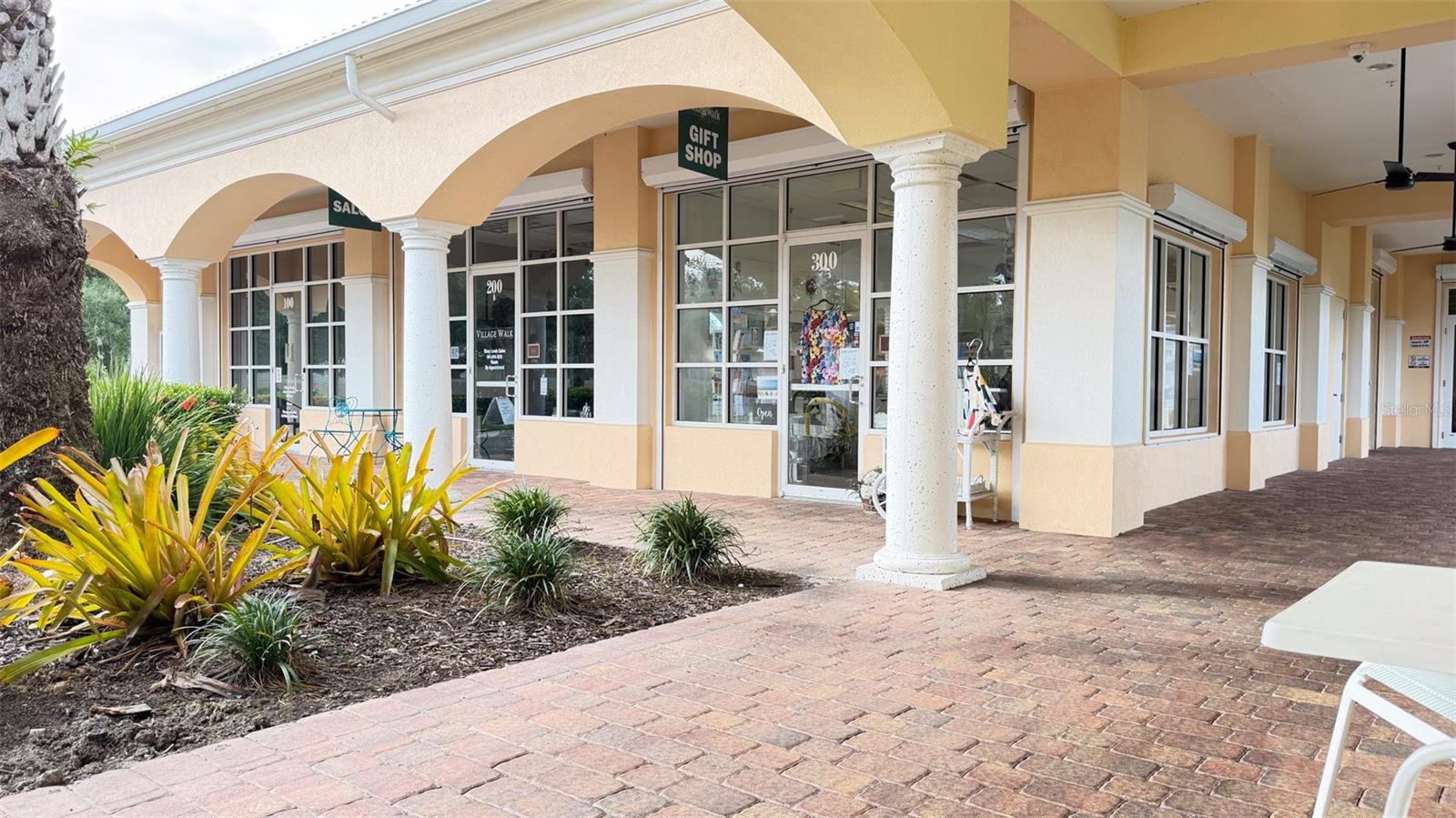 Village Walk storefronts beneath cream arches, with white columns and a brick-paved walkway.