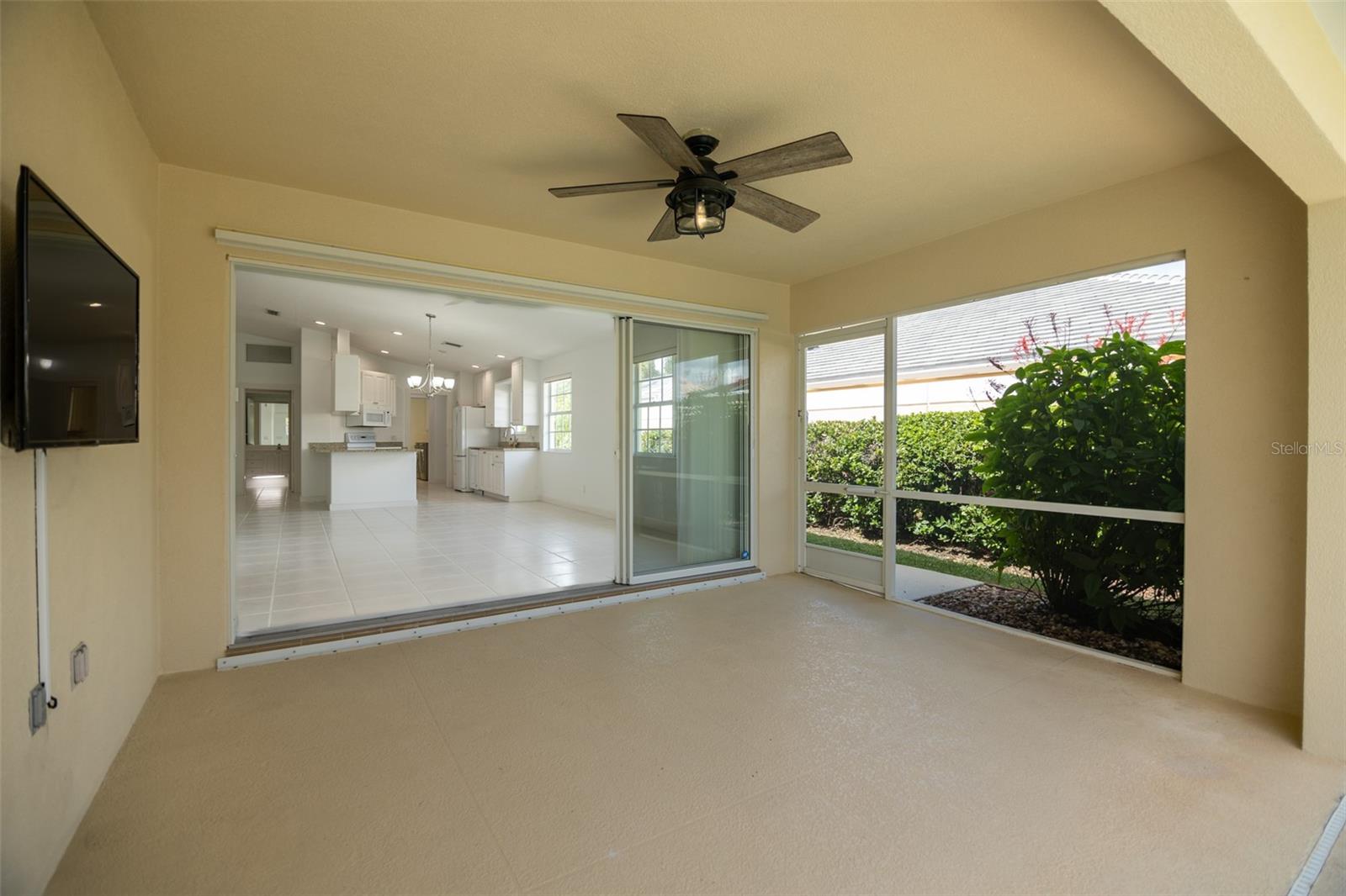 Covered lanai with ceiling fan, sliding doors to a white kitchen, and screened windows overlooking shrubs.