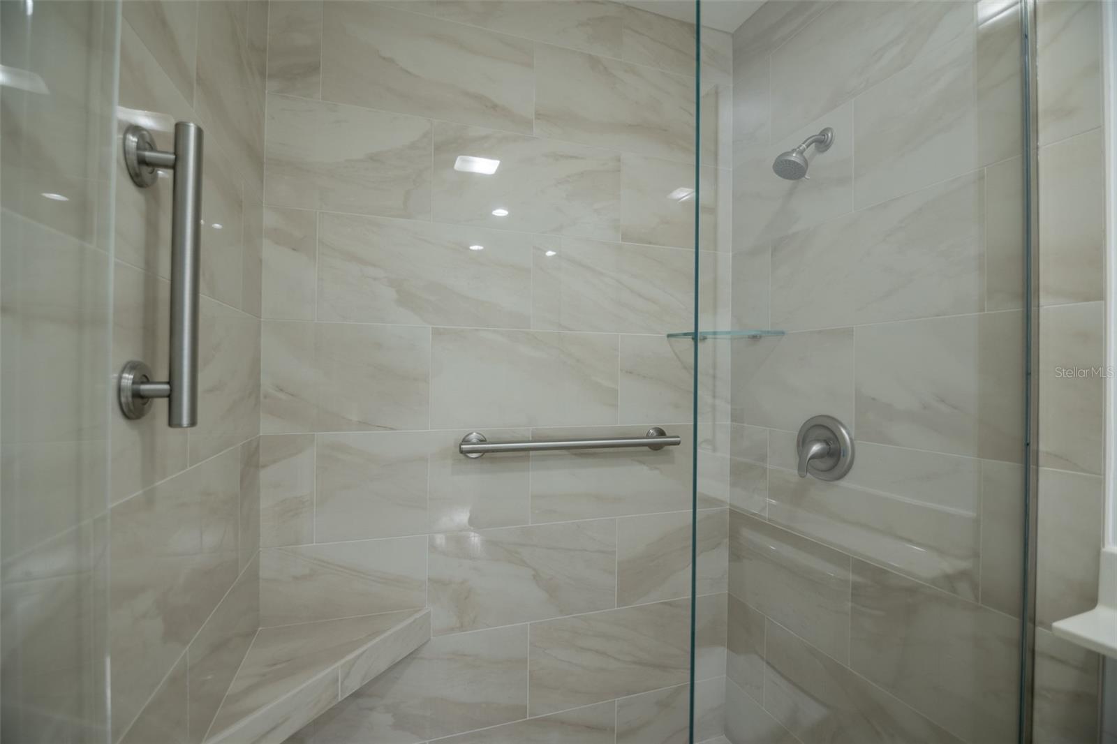 Walk-in shower with cream marble-look tile, glass enclosure, metal grab bars, and a built-in bench.