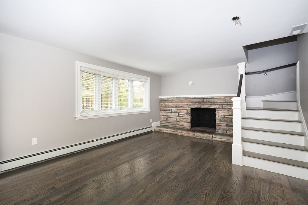 Empty living room with a stone fireplace, dark hardwood floors, and stairs.