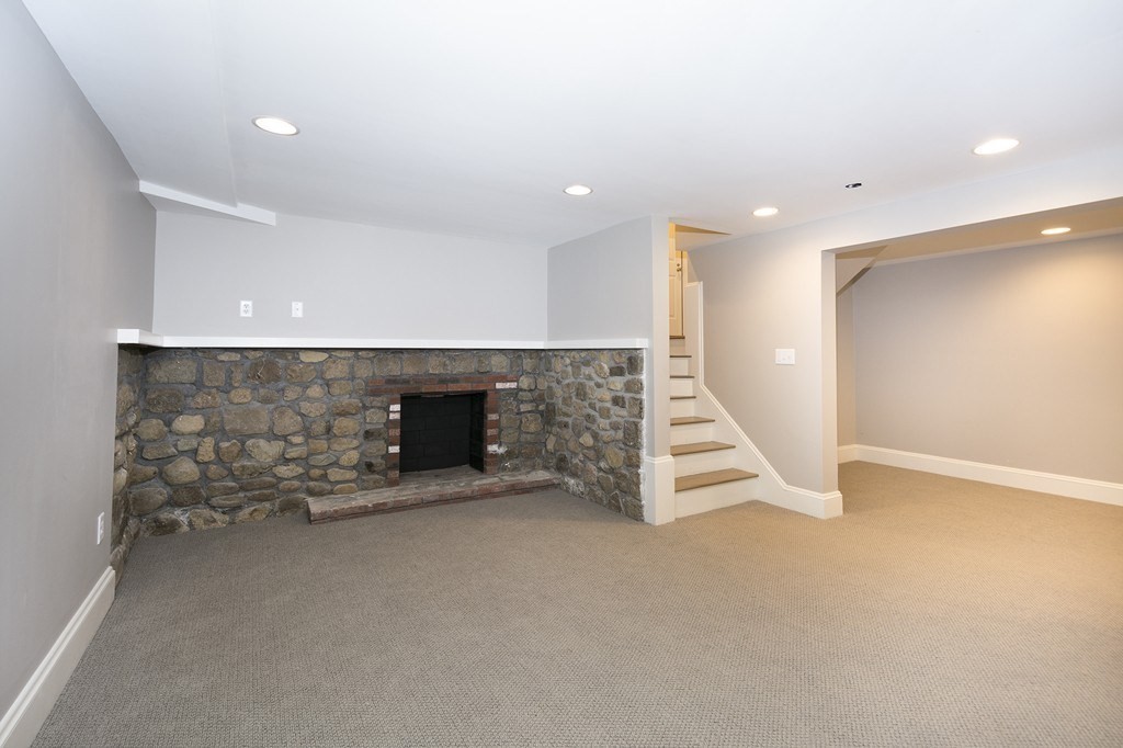 Empty basement living area with a stone fireplace and stairs.